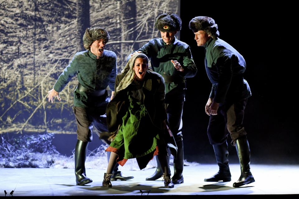 A group of students, in dark blue and green clothes, performing on stage, surrounded by a dark stage setting.